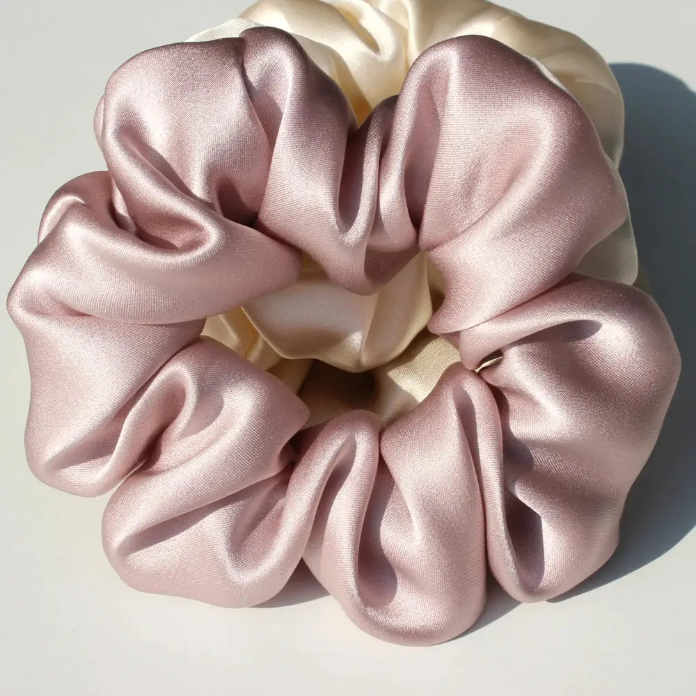 Hand holding two large 100% pure mulberry silk scrunchies in blush pink and ivory. Soft and smooth silk hair ties for a stylish, damage-free hold.