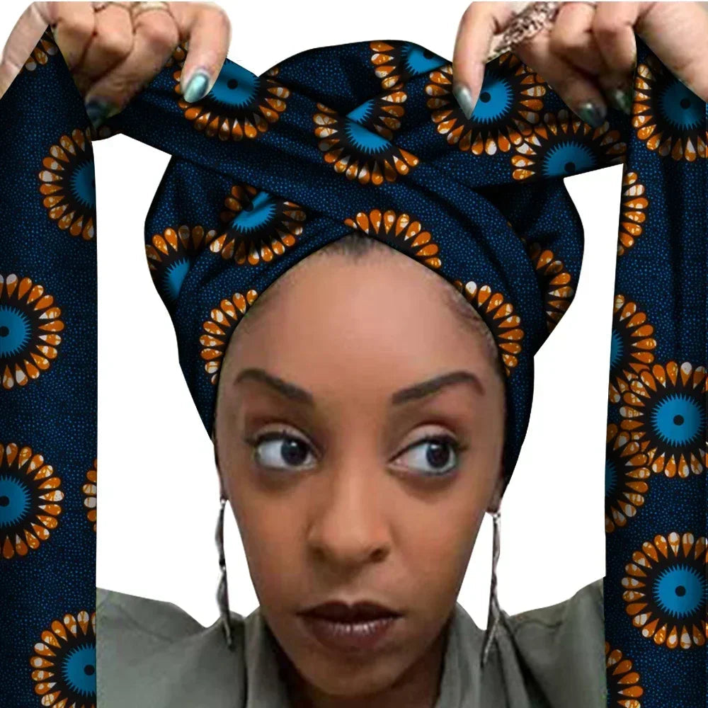 Vrouw met een Afrikaanse headwrap in donkerblauw en oranje Ankara print met bloemachtig zonvormig patroon. De stof heeft levendige kleuren en lange linten, handgemaakt van katoen, perfect voor lente en zomer.