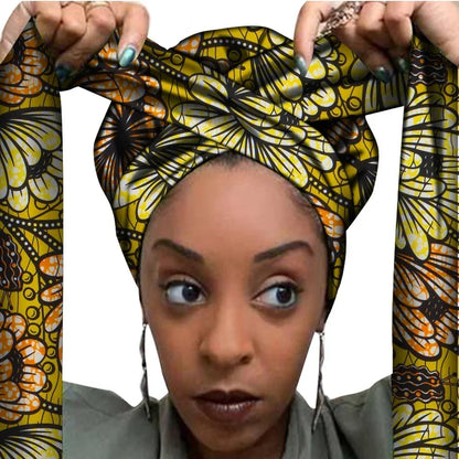 Vrouw met een Afrikaanse headwrap in geel, zwart en wit Ankara print met oranje accenten. De stof heeft een bloemachtig patroon met elegante lijnen en vormen, lange linten en is handgemaakt van katoen, perfect voor lente en zomer.