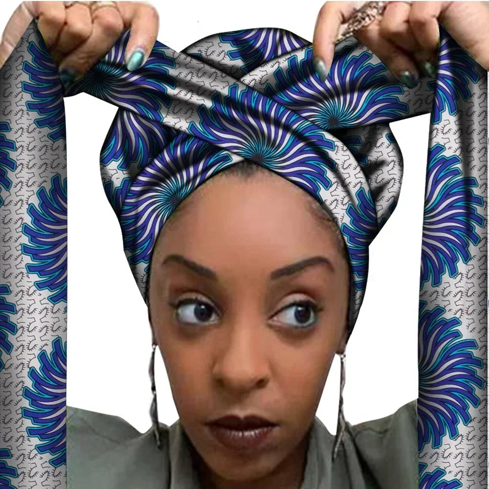 Vrouw met een Afrikaanse headwrap in grijs, blauw en paars Ankara print. De stof heeft een bloemachtig patroon met stralende lijnen en decoratieve details, lange linten en is handgemaakt van katoen, perfect voor lente en zomer