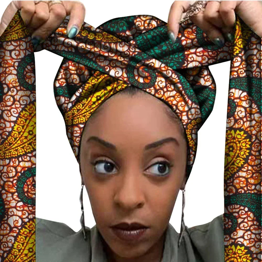 Vrouw met een Afrikaanse headwrap in groen, geel en oranje Ankara print met bruine details. De stof heeft een sierlijk, gedetailleerd patroon met krullen en bloemachtige vormen, lange linten en is handgemaakt van katoen, perfect voor lente en zomer.