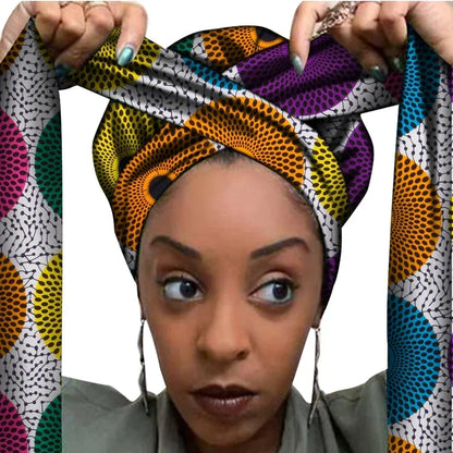 Vrouw met een Afrikaanse headwrap in een kleurrijke Ankara print met geel, paars, oranje, groen, blauw en wit. De stof heeft een opvallend cirkelvormig patroon met stippen en lange linten, handgemaakt van katoen, perfect voor lente en zomer.