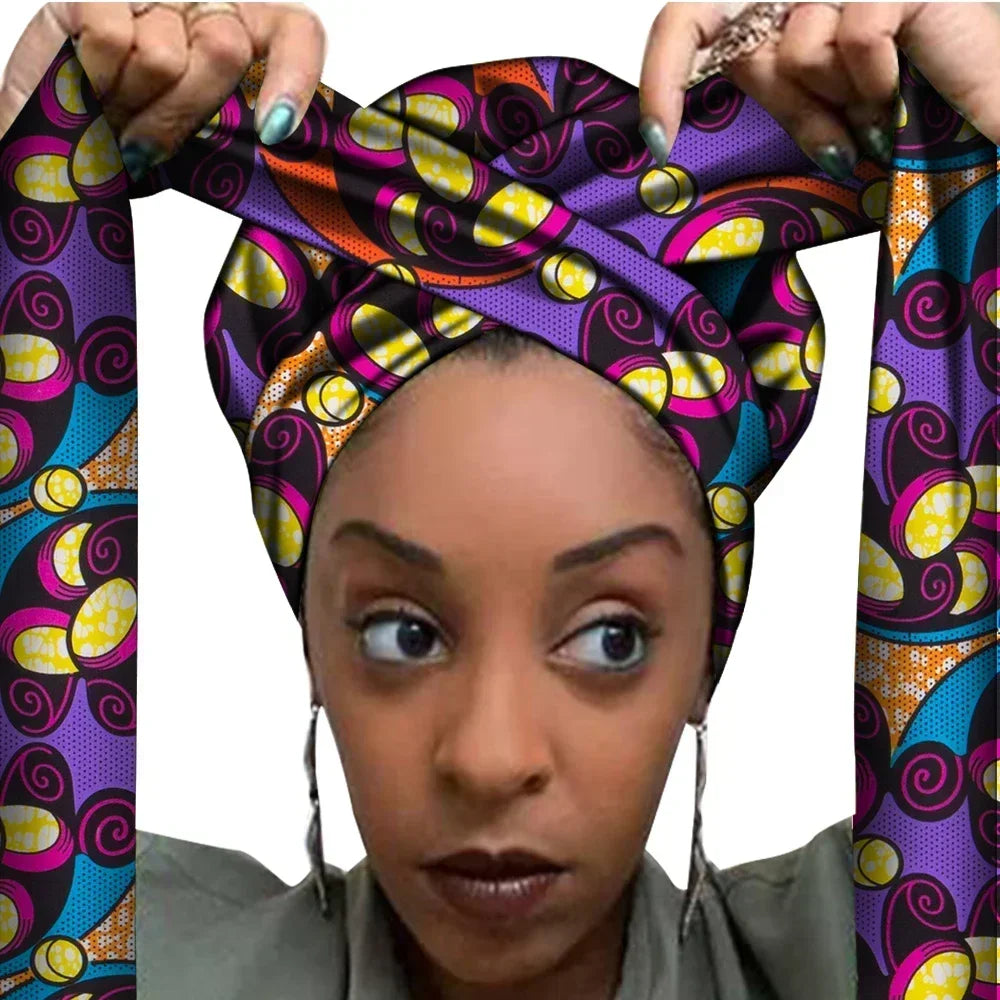 Vrouw met een Afrikaanse headwrap in paars, roze, geel en zwart Ankara print. De stof heeft een speels, golvend patroon met kleurrijke accenten en lange linten, handgemaakt van katoen, ideaal voor lente en zomer.
