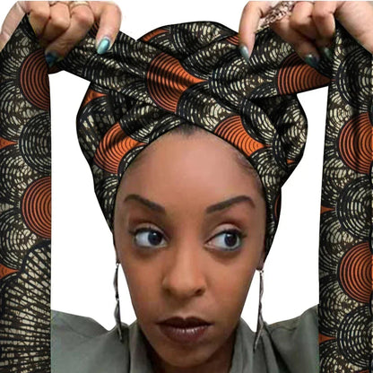 Vrouw met een Afrikaanse headwrap in zwart en oranje Ankara print met beige details. De stof heeft een cirkelvormig en grafisch patroon met lange linten, handgemaakt van katoen, ideaal voor lente en zomer.