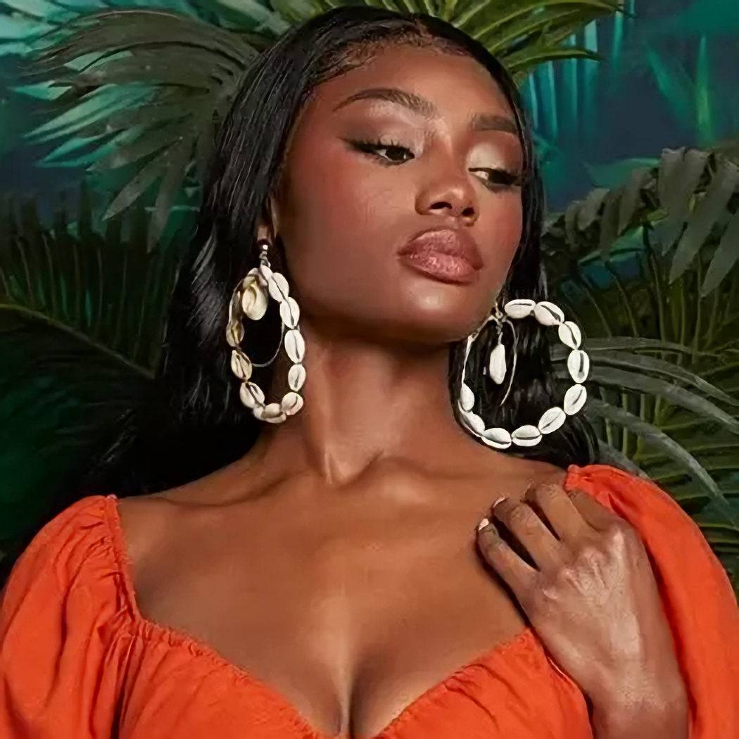 Model wearing large double hoop Cowrie Shell earrings, styled with a tropical backdrop and an orange off-shoulder dress.