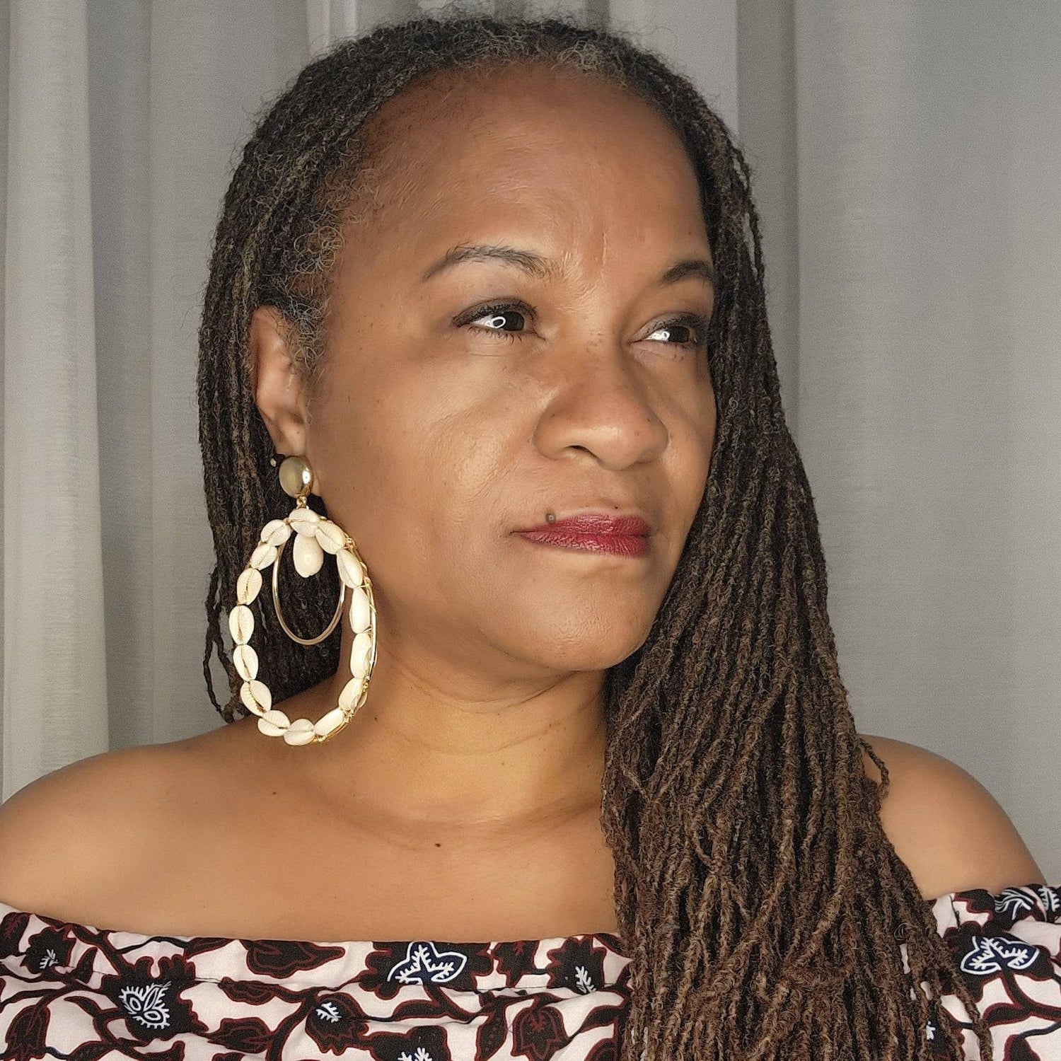 Woman with long dreadlocks wearing statement double hoop Cowrie Shell earrings, paired with an off-shoulder printed top.