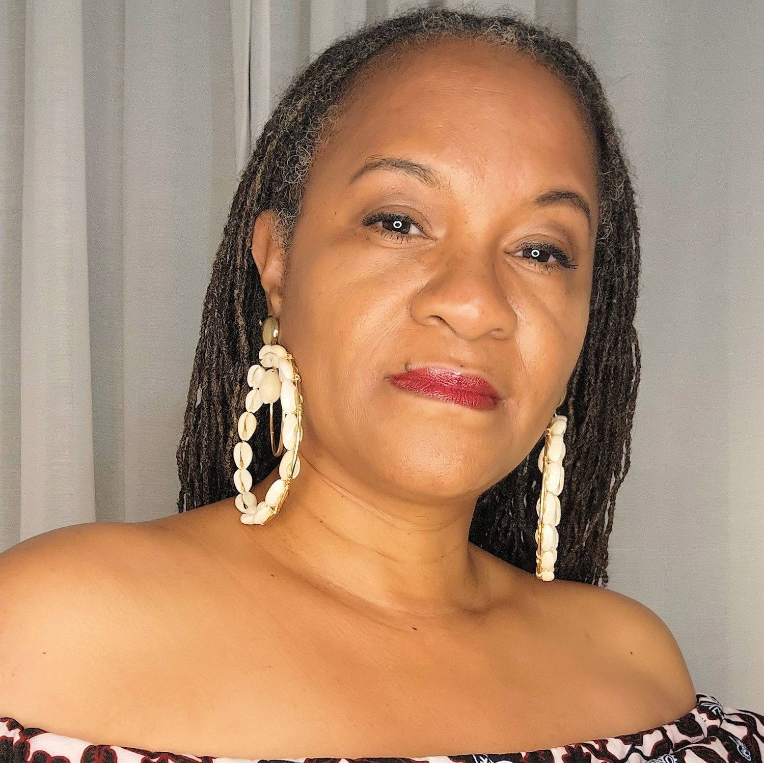 Woman wearing double hoop Cowrie Shell earrings with gold-tone accents, styled with long dreadlocks and an off-shoulder top.